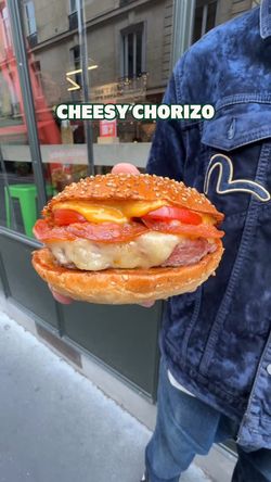 Burger avec fromage, chorizo, tomates et pain sésame dans un environnement extérieur.