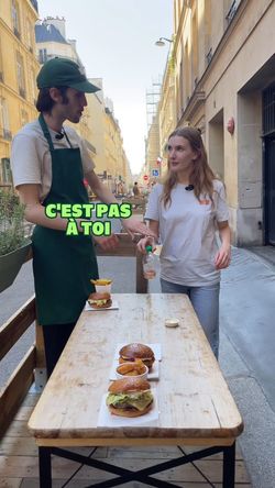Deux burgers bio et un burger vegan sur une table à l'extérieur, avec une serveuse et un serveur en discussion.