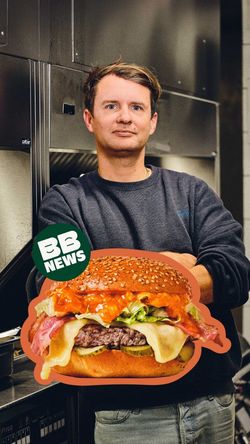 Un homme tient un burger bio garni de fromage, bacon, salade et sauce, dans un restaurant burger bio convivial.