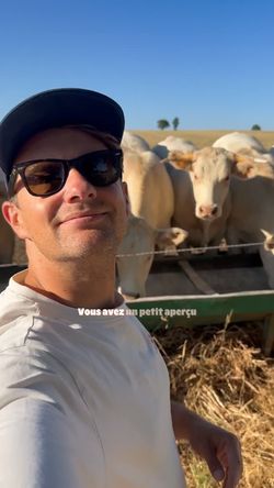 Un homme portant une casquette et des lunettes de soleil prend un selfie devant un troupeau de vaches dans un champ sous un C