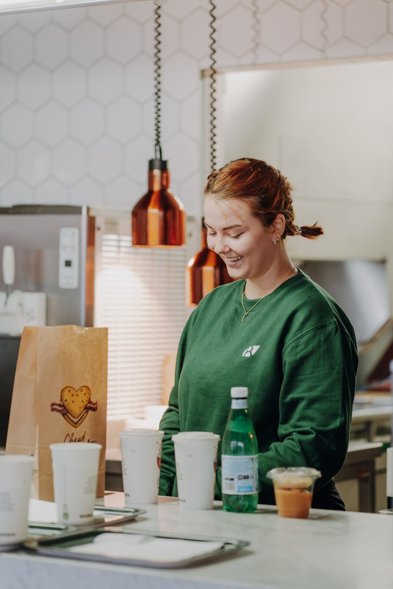 Une employée souriante prépare des commandes avec des boissons et un sac en papier sur le comptoir.
