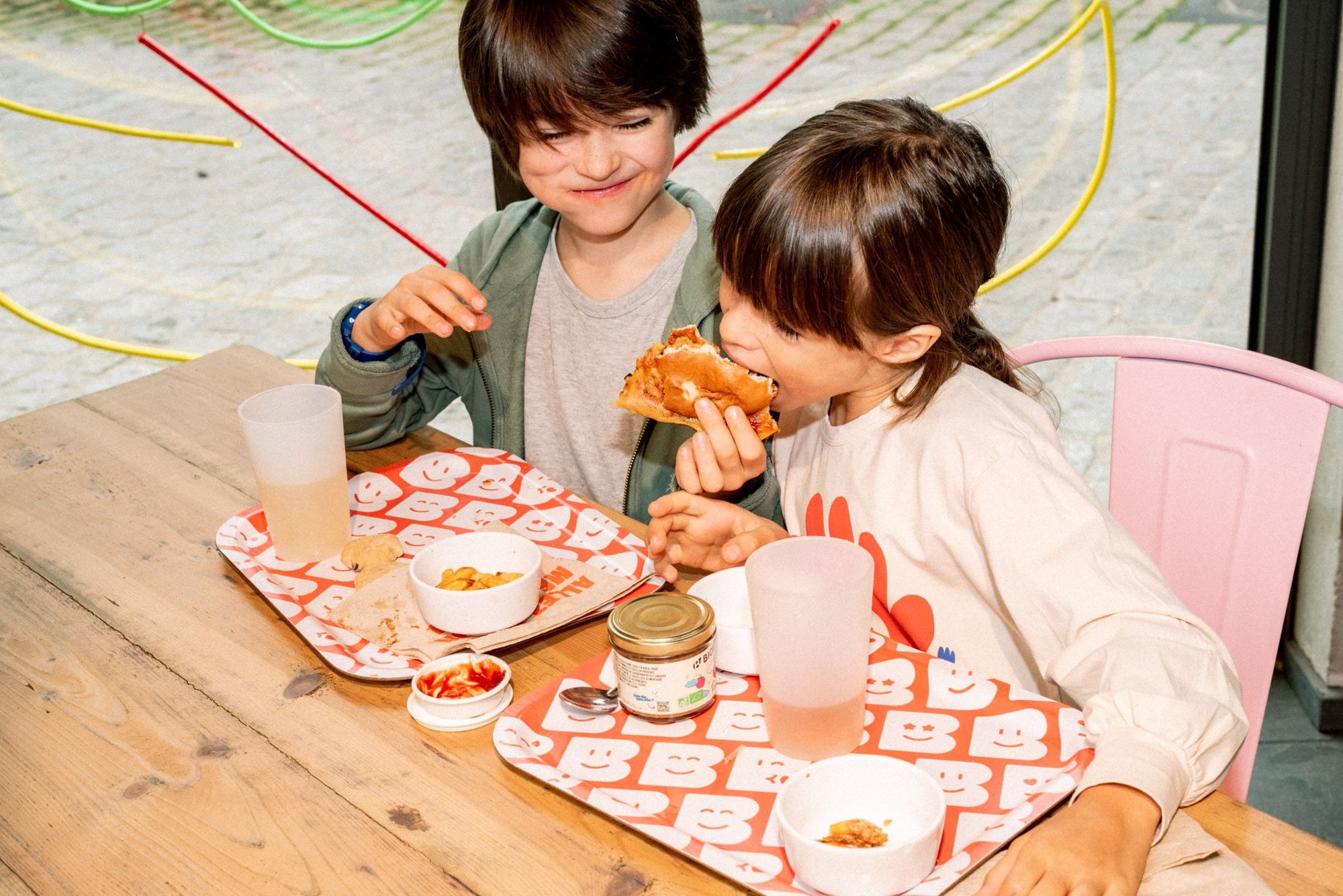 Deux enfants dégustent des burgers bio sur une table en bois, avec des boissons et des accompagnements.