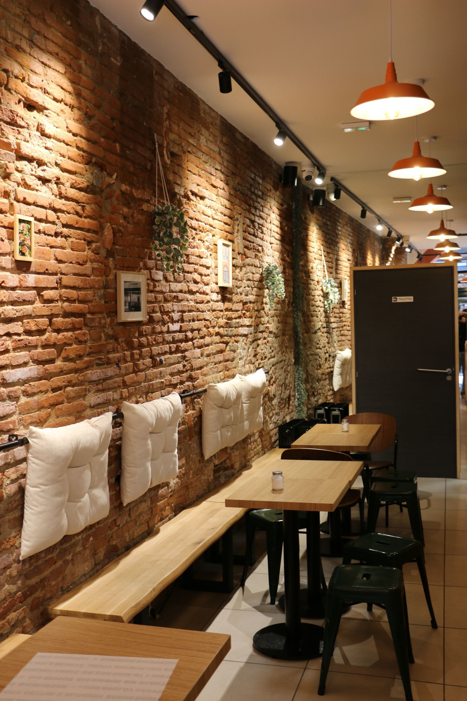 Intérieur du restaurant bio avec murs en brique, banquette en bois et coussins blancs, tables et chaises noires modernes.