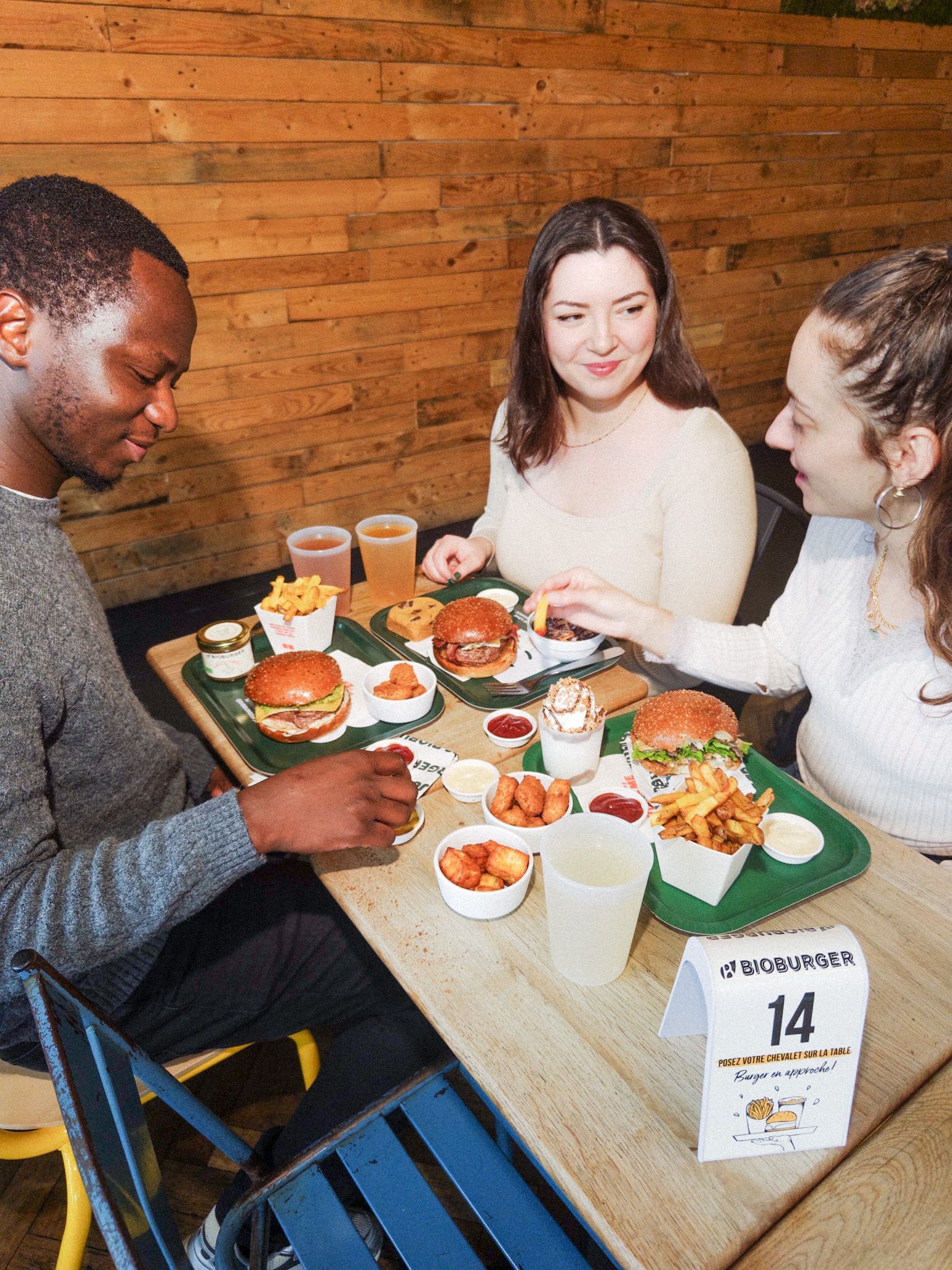 Trois personnes dégustent un repas avec des burgers bio, frites, et boissons dans une ambiance décontractée.