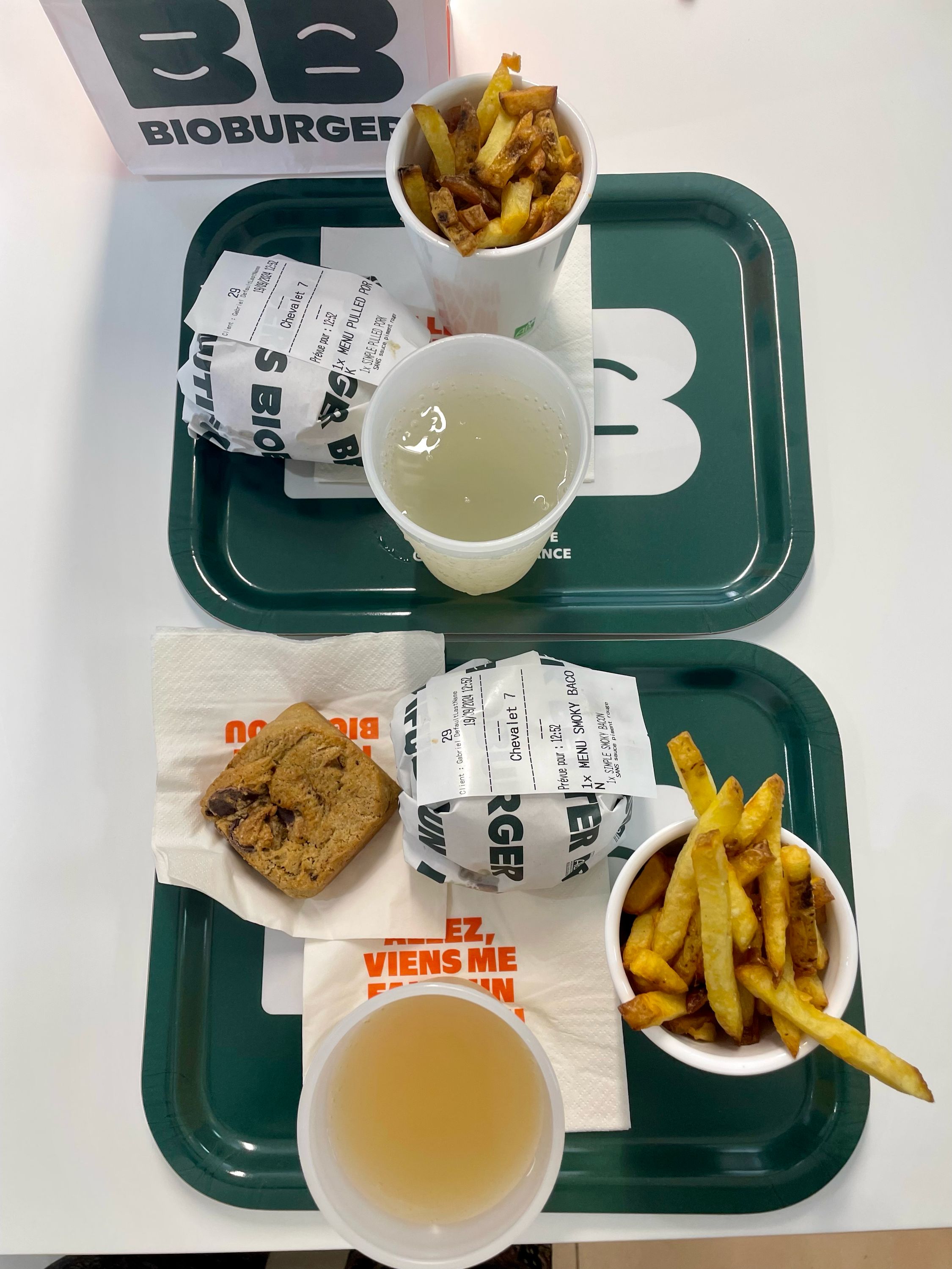 Deux plateaux avec des burgers bio, frites et boissons, incluant un cookie en dessert.