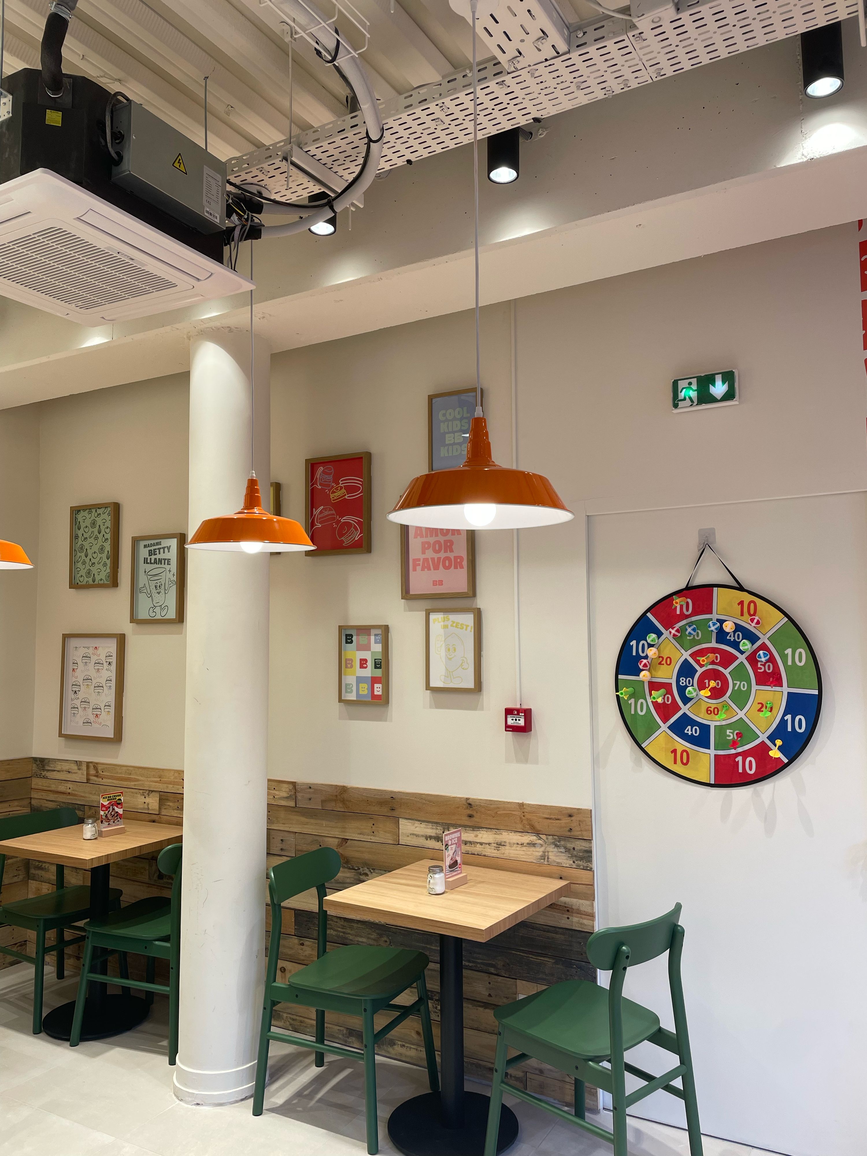 Intérieur d'un restaurant avec tables en bois, chaises vertes, lampes suspendues orange, et décoration murale.