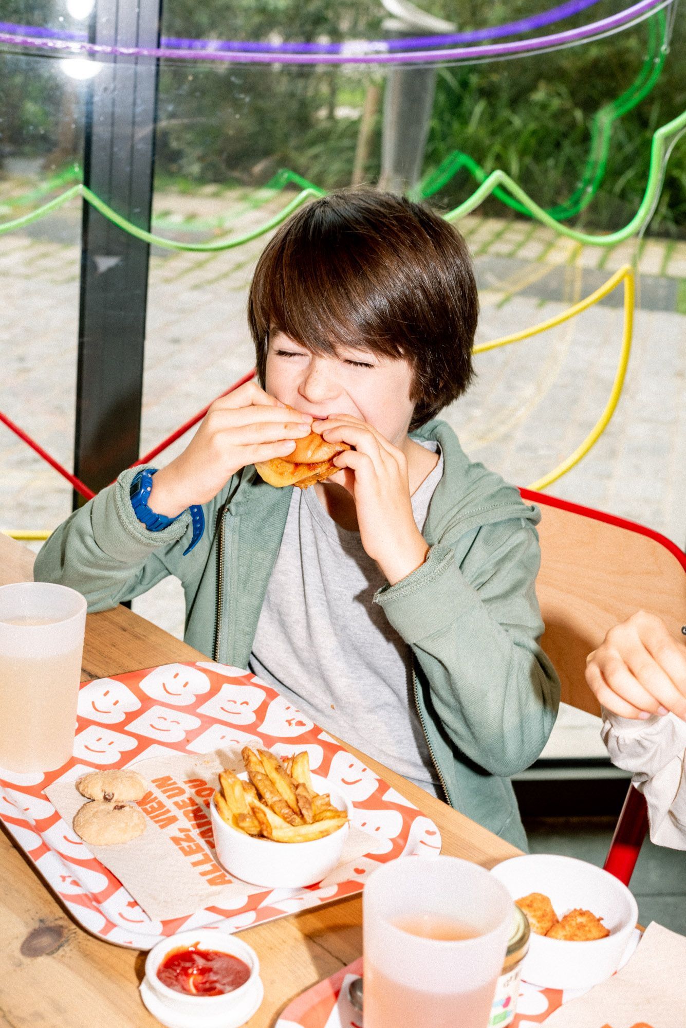 Un enfant souriant déguste un burger avec des frites, assis à une table dans un cadre coloré.