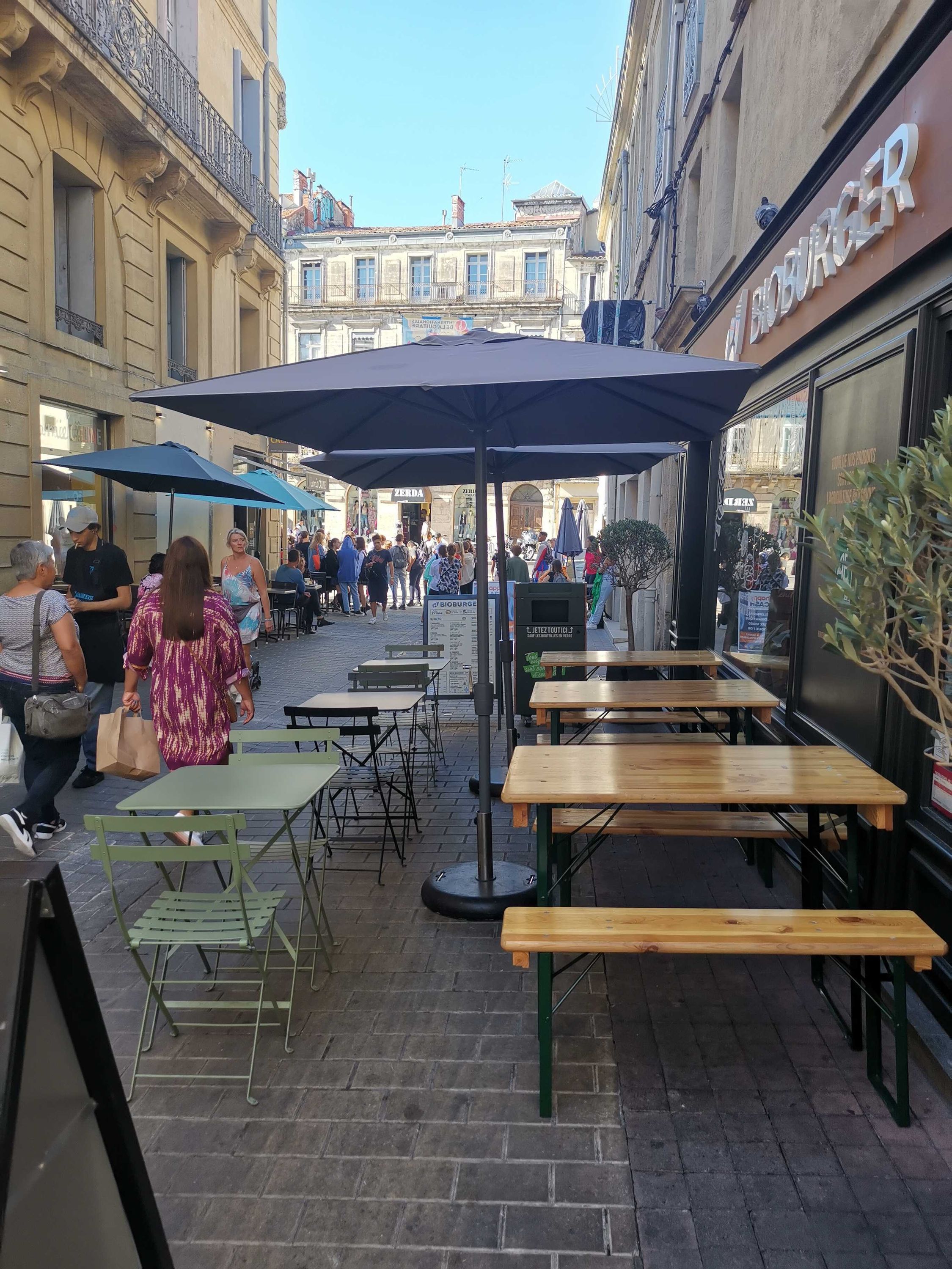 Terrasse avec tables en bois et parasols, située dans une rue animée près du Bioburger Montpellier.