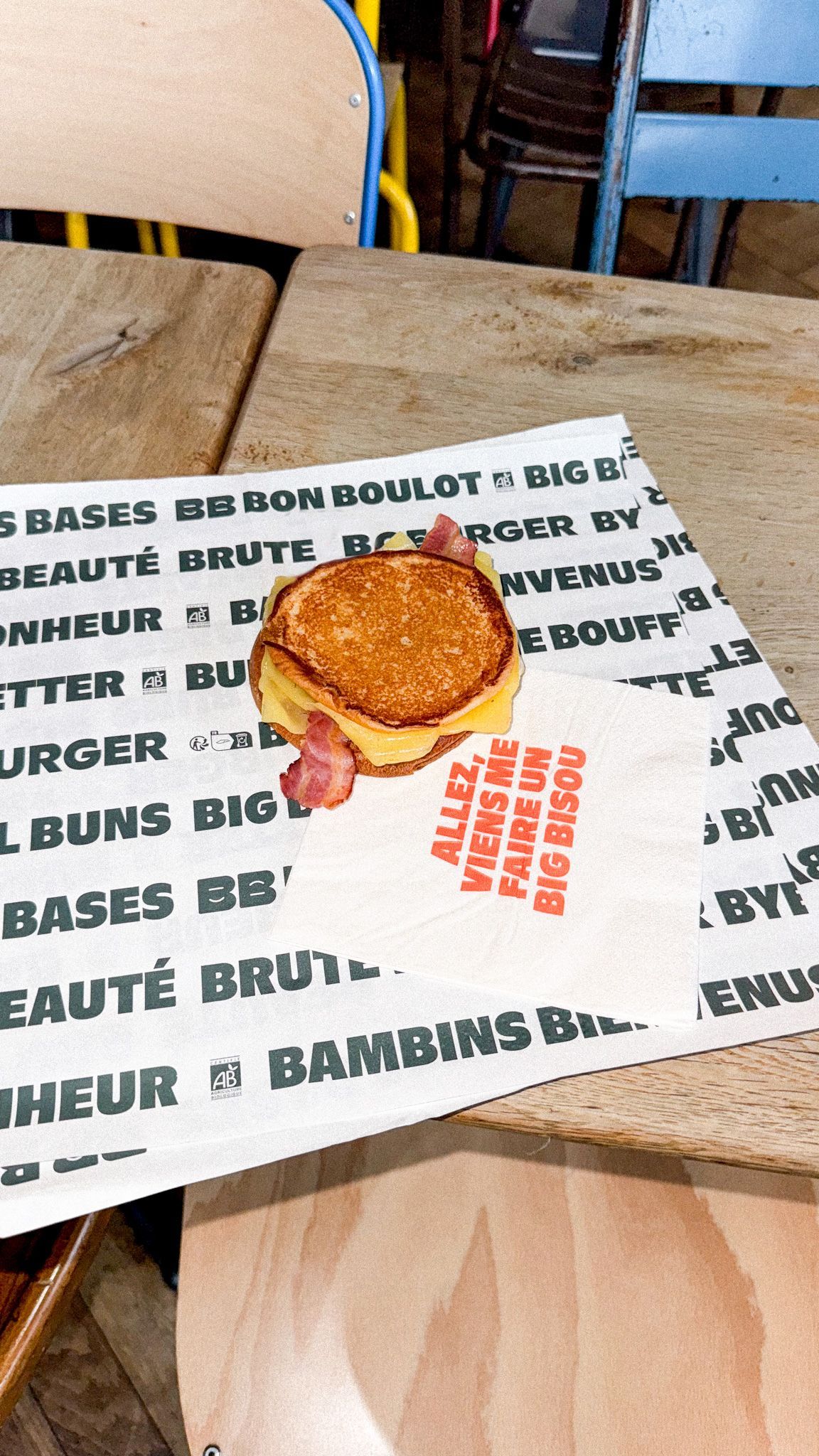 Un burger au bacon présenté sur du papier imprimé, accompagné de chaises colorées en arrière-plan.