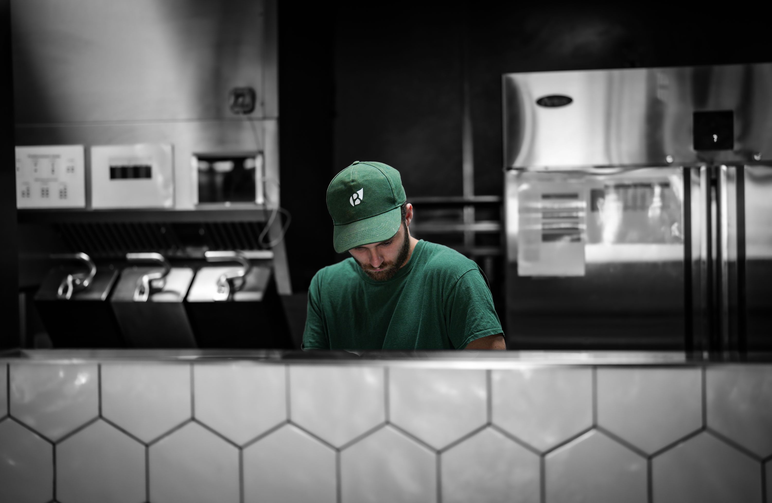 Un cuisinier prépare des plats dans la cuisine d'un restaurant, portant une tenue verte et une casquette.