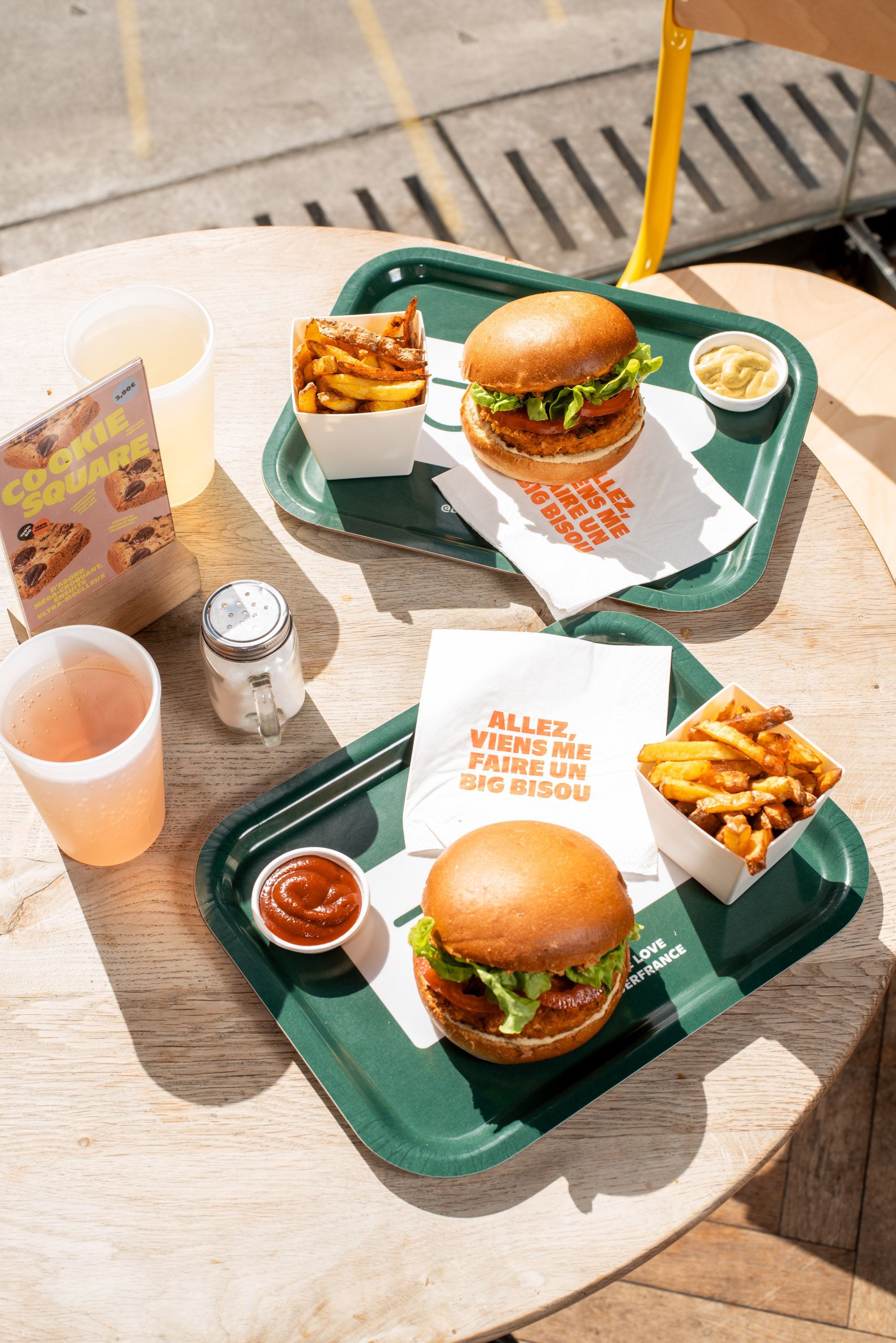 Deux plateaux avec burgers bio, frites et sauces, sur une table en bois ensoleillée.