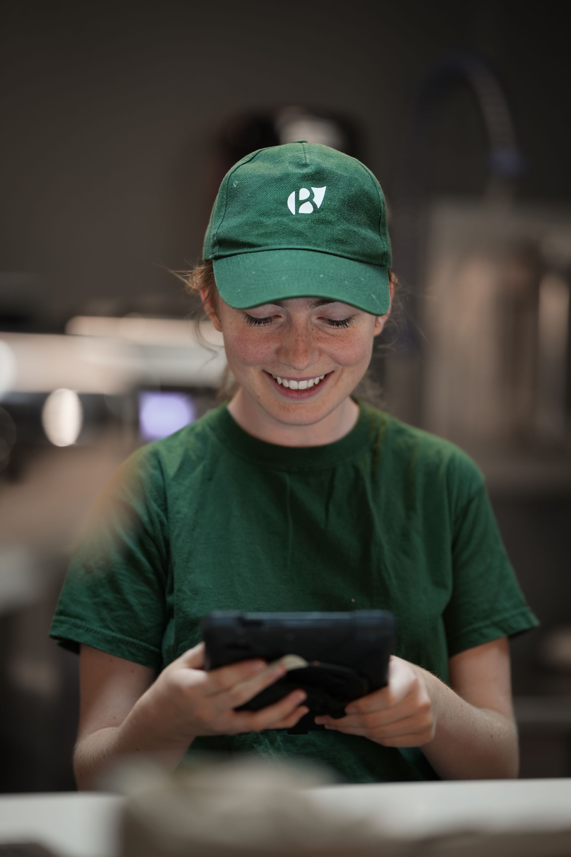 Employé souriant avec un t-shirt vert et une casquette, utilisant une tablette dans un restaurant.